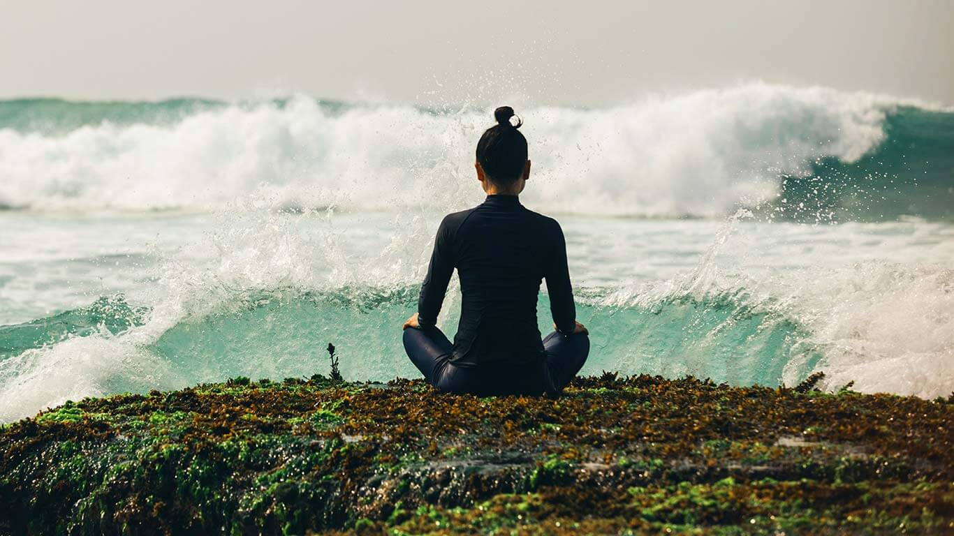 Young woman in yoga pose sitting on beach, with her back to the camera, watching the surf come in.